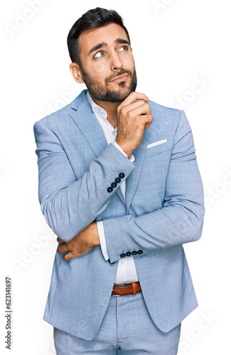 Young hispanic man wearing business jacket serious face thinking about question with hand on chin, thoughtful about confusing idea