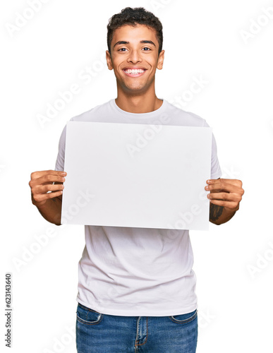 Young handsome african american man holding blank empty banner looking positive and happy standing and smiling with a confident smile showing teeth