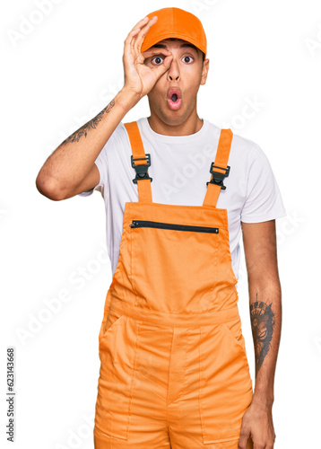 Young handsome african american man wearing handyman uniform doing ok gesture shocked with surprised face, eye looking through fingers. unbelieving expression.