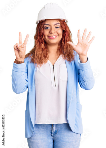 Young latin woman wearing architect hardhat showing and pointing up with fingers number seven while smiling confident and happy.