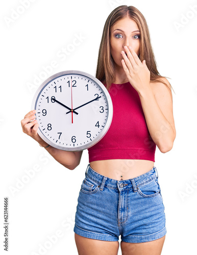 Young beautiful blonde woman holding big clock covering mouth with hand, shocked and afraid for mistake. surprised expression