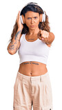 Young hispanic woman with tattoo listening to music using headphones pointing with finger to the camera and to you, confident gesture looking serious