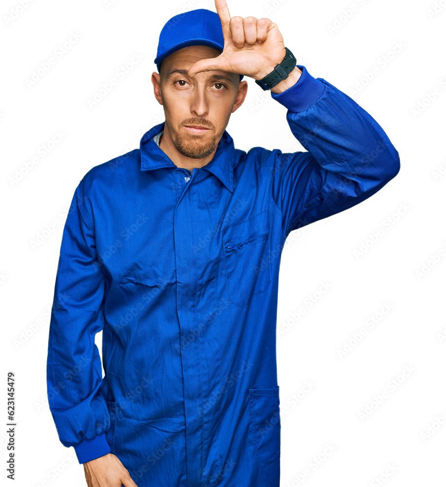 Bald man with beard wearing builder jumpsuit uniform making fun of people with fingers on forehead doing loser gesture mocking and insulting.
