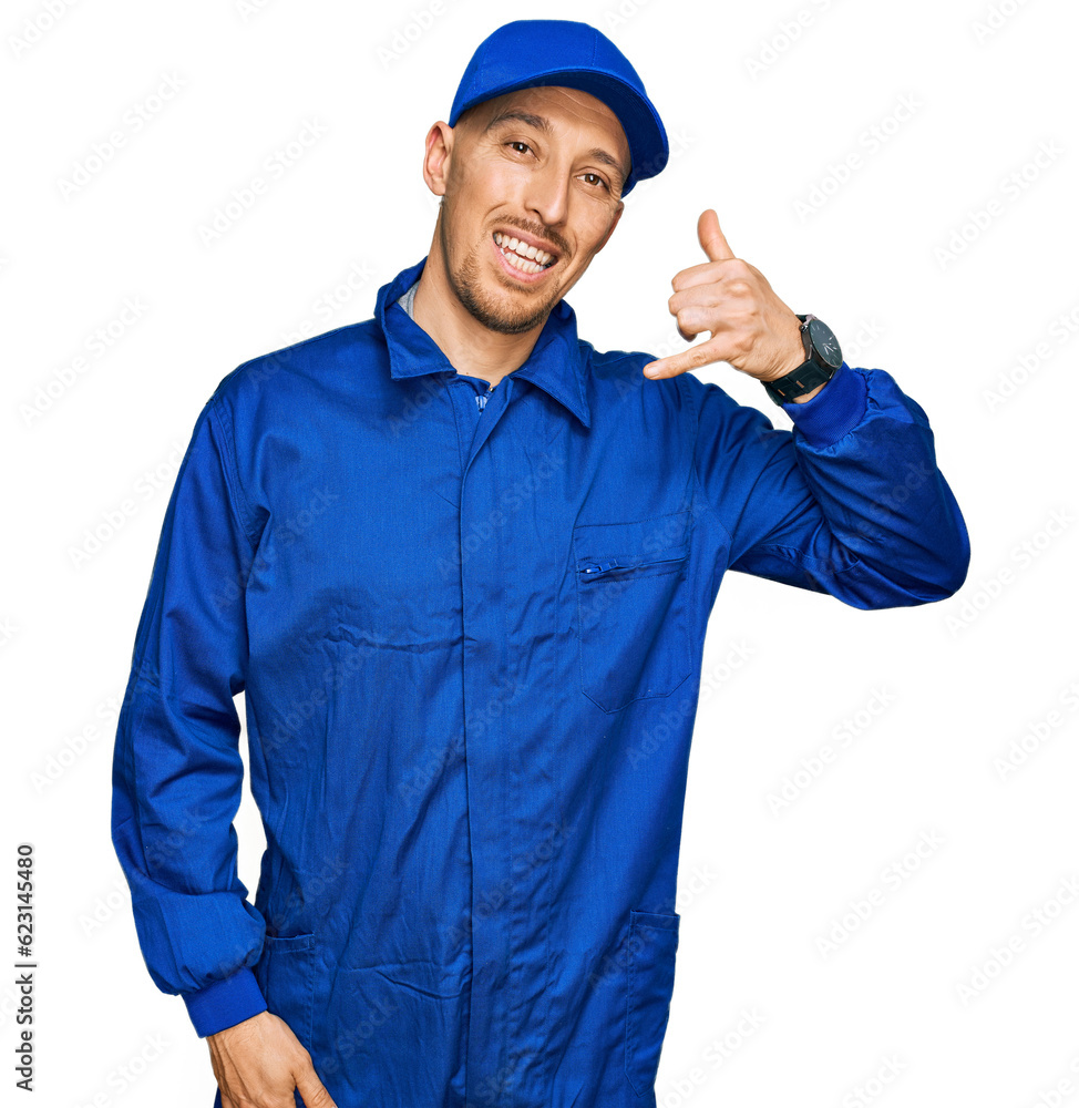 Bald man with beard wearing builder jumpsuit uniform smiling doing phone gesture with hand and fingers like talking on the telephone. communicating concepts.