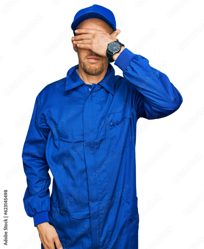 Bald man with beard wearing builder jumpsuit uniform covering eyes with hand, looking serious and sad. sightless, hiding and rejection concept