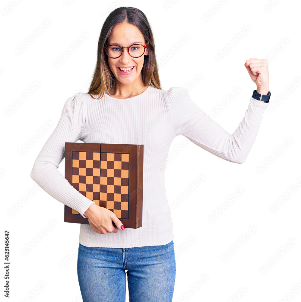 Beautiful brunette young woman holding chess screaming proud, celebrating victory and success very excited with raised arms Beautiful brunette young woman holding chess screaming proud, celebrating victory and success very excited with raised arms