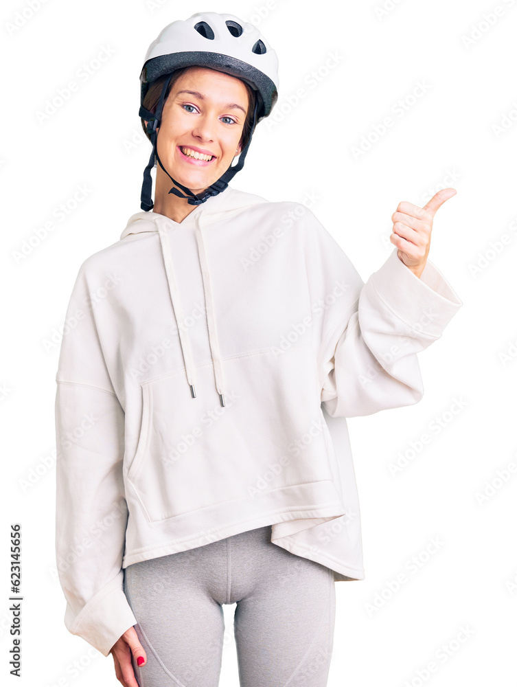 Beautiful brunette young woman wearing bike helmet and sporty clothes smiling with happy face looking and pointing to the side with thumb up. Beautiful brunette young woman wearing bike helmet and sporty clothes smiling with happy face looking and pointing to the side with thumb up.