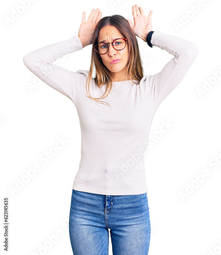 Beautiful brunette young woman wearing casual white sweater and glasses doing bunny ears gesture with hands palms looking cynical and skeptical. easter rabbit concept. photo