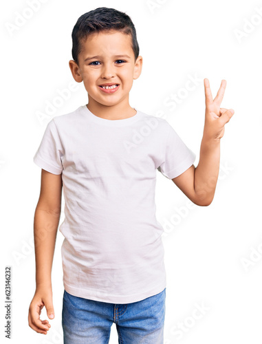 Little cute boy kid wearing casual white tshirt smiling with happy face winking at the camera doing victory sign. number two.