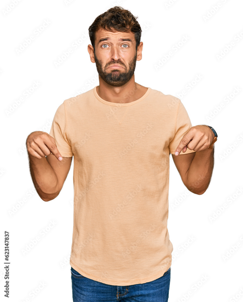 Handsome young man with beard wearing casual tshirt pointing down looking sad and upset, indicating direction with fingers, unhappy and depressed. Handsome young man with beard wearing casual tshirt pointing down looking sad and upset, indicating direction with fingers, unhappy and depressed.