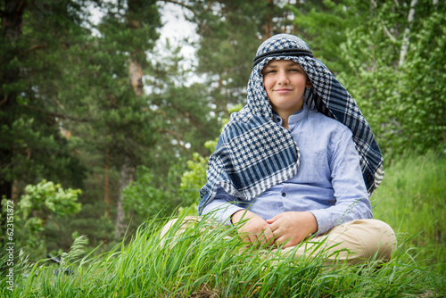 In summer, a boy in an Arab scarf sits on the grass.