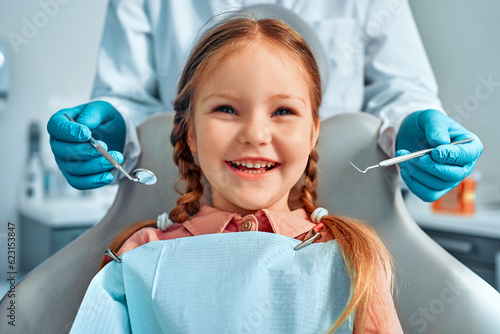 Cropped portrait of girl with pigtails hair sitting in dental chair looking at camera and smiling. Behind  a doctor in gloves holds examination tools.Children s dentistry.