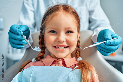 Cropped portrait of little girl with pigtails hair sitting in dental chair looking at camera and smiling. Behind, a doctor in gloves holds examination tools.Children's dentistry.