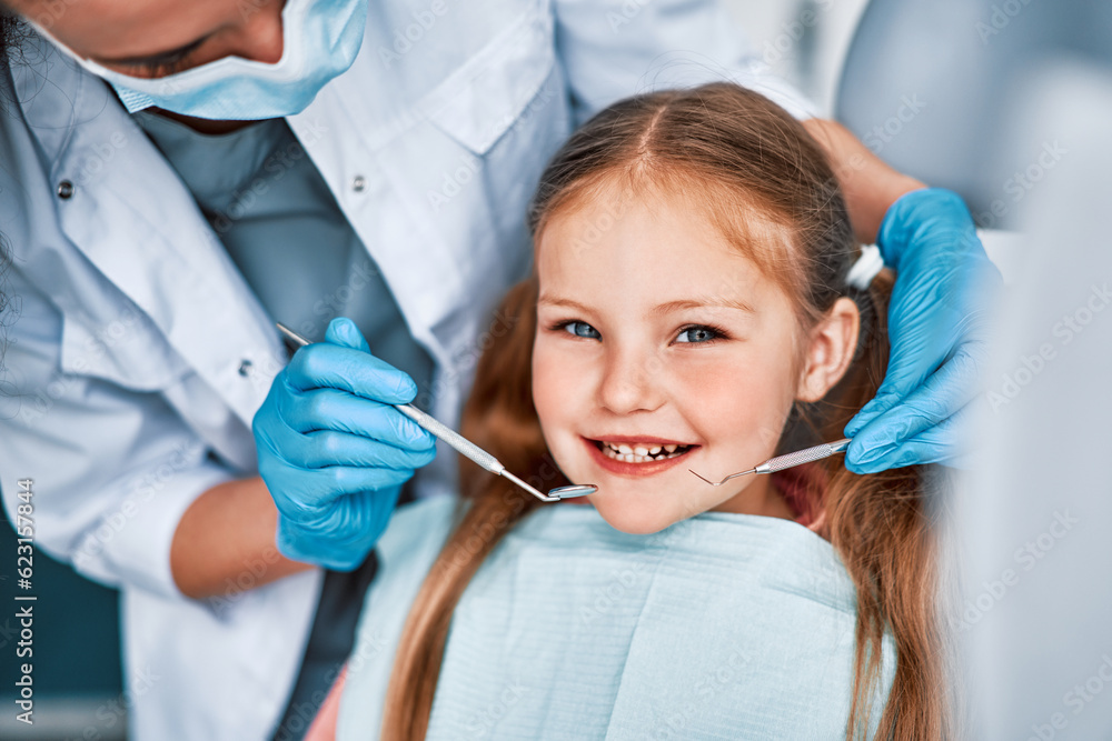 Photo of a child smiling at a dentist appointment. A doctor in gloves holds examination tools behind. Children's dentistry. Photo of a child smiling at a dentist appointment. A doctor in gloves holds examination tools behind. Children's dentistry.