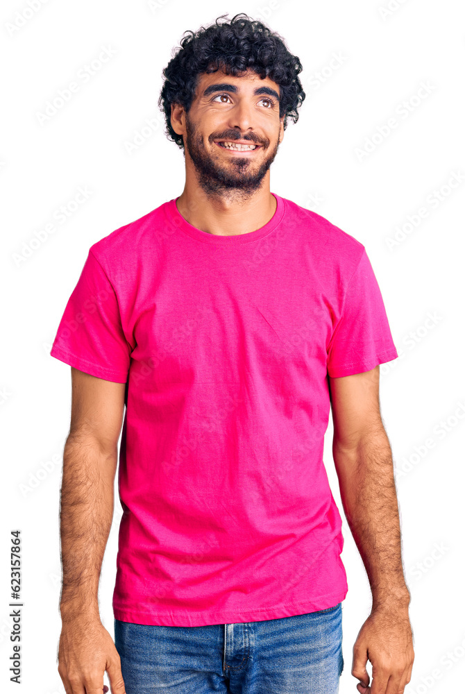 Handsome young man with curly hair and bear wearing casual pink tshirt looking away to side with smile on face, natural expression. laughing confident. Handsome young man with curly hair and bear wearing casual pink tshirt looking away to side with smile on face, natural expression. laughing confident.