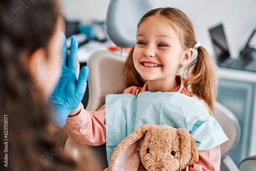 At the doctor's appointment. A candid emotional photo of a child sitting in a dental chair, holding a toy rabbit and cheerfully giving a high-five to the nurse. photo