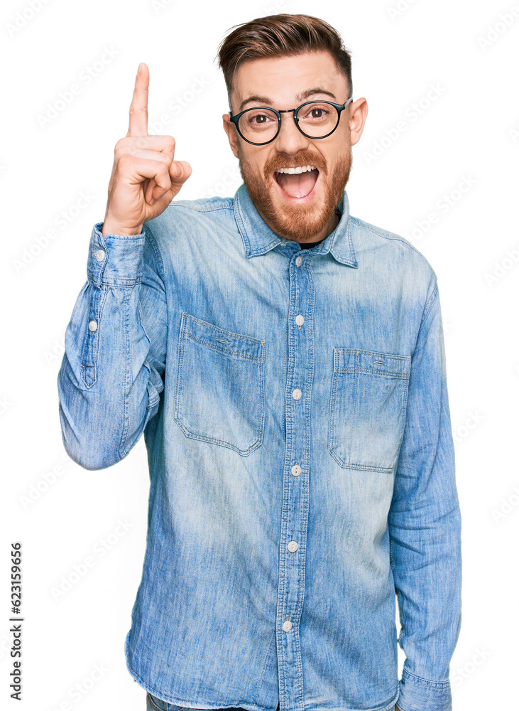 Young redhead man wearing casual denim shirt pointing finger up with successful idea. exited and happy. number one. Young redhead man wearing casual denim shirt pointing finger up with successful idea. exited and happy. number one.