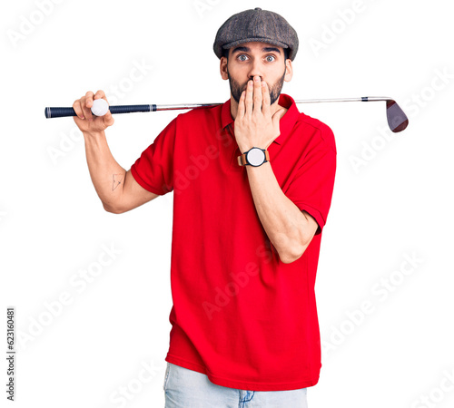 Young handsome man with beard playing golf holding club and ball covering mouth with hand, shocked and afraid for mistake. surprised expression