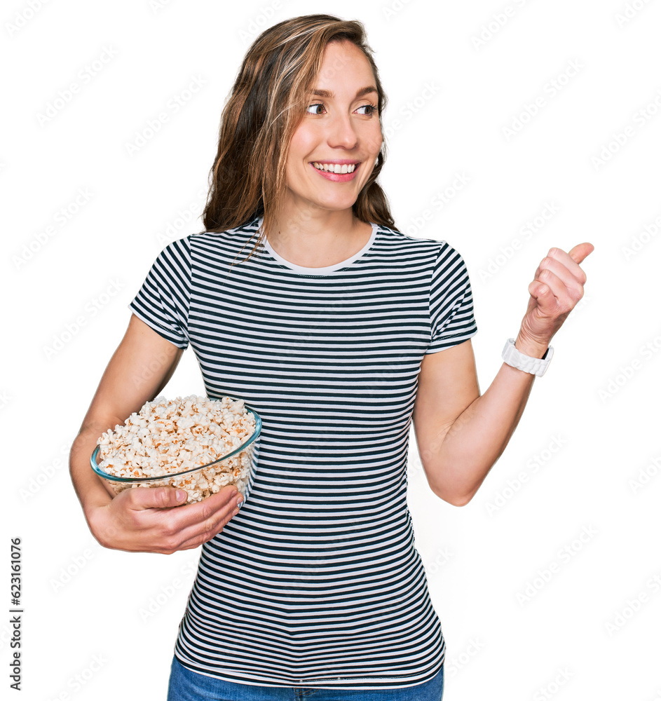 Young blonde woman eating popcorn pointing thumb up to the side smiling happy with open mouth Young blonde woman eating popcorn pointing thumb up to the side smiling happy with open mouth