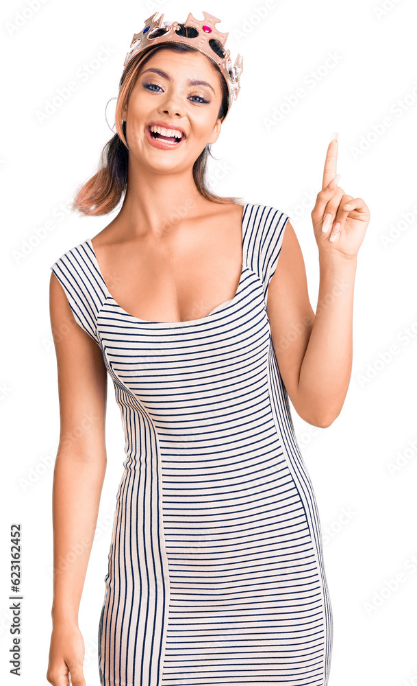 Young beautiful woman wearing king crown pointing finger up with successful idea. exited and happy. number one. Young beautiful woman wearing king crown pointing finger up with successful idea. exited and happy. number one.