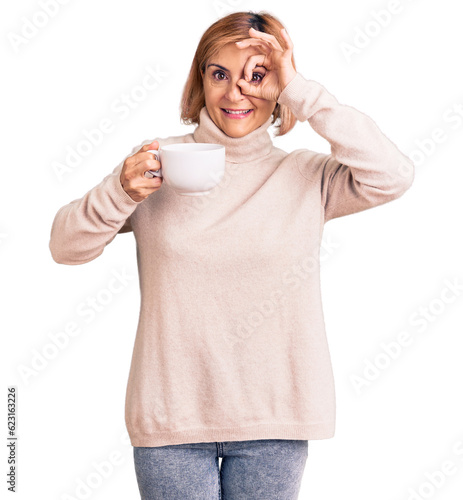 Young blonde woman holding coffee smiling happy doing ok sign with hand on eye looking through fingers