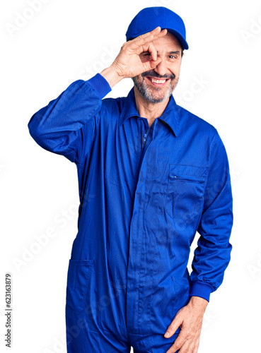Middle age handsome man wearing mechanic uniform doing ok gesture with hand smiling, eye looking through fingers with happy face.