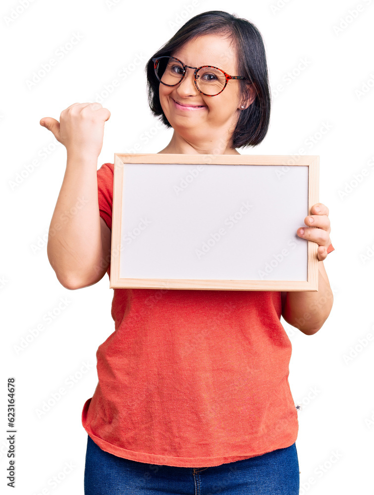Brunette woman with down syndrome holding empty white chalkboard pointing thumb up to the side smiling happy with open mouth Brunette woman with down syndrome holding empty white chalkboard pointing thumb up to the side smiling happy with open mouth