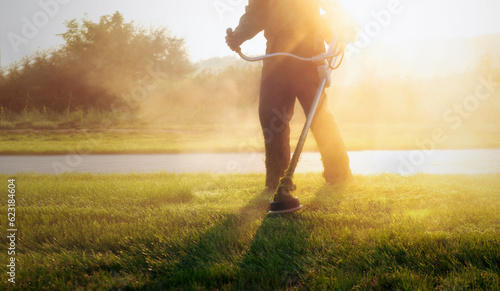 Close-up shot of municipal worker holding a lawnmower, trimming the grass in a public park during a vibrant sunrise. Maintaining the lawn at dawn with dramatic sunshine. photo