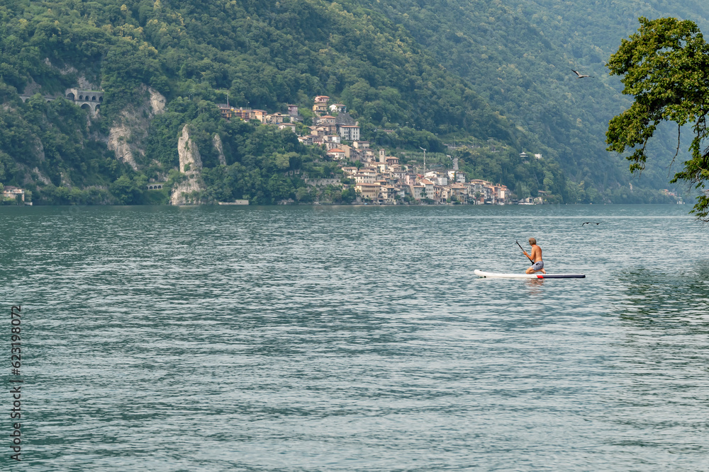 A man kneeling on a stand up paddle on Lake Lugano, Switzerland, with Gandria in the background
