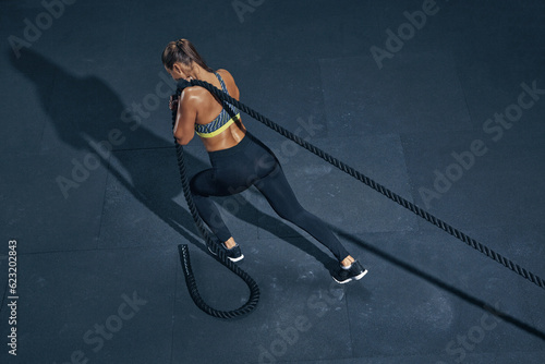 Female athlete fitness rope pull training. Sporty sportswoman working out in functional training gym doing cross exercise with battle rope