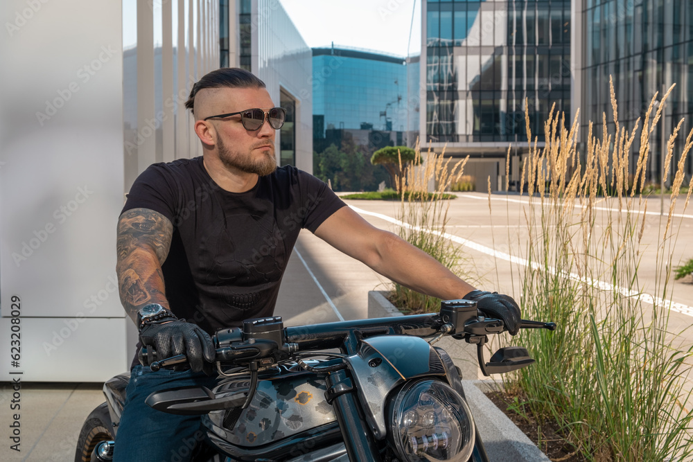 Handsome motorcyclist in sunglasses on his moto against urban backdrop in city