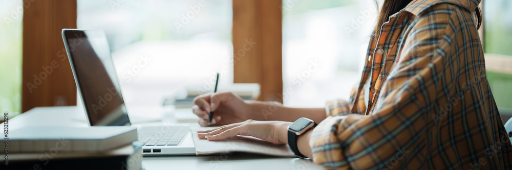 Fototapeta premium Young woman student busy study make notes in notebook and using laptop concentrated online training course technology education.