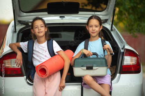 Two happy twin sisters with backpacks with a tent in their hands put tourist things in the trunk of a white car for going outdoors for recreation