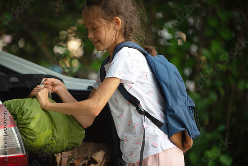 a happy girl in a white T-shirt with a backpack with a tent in her hands puts in the trunk of a white car for going outdoors for recreation