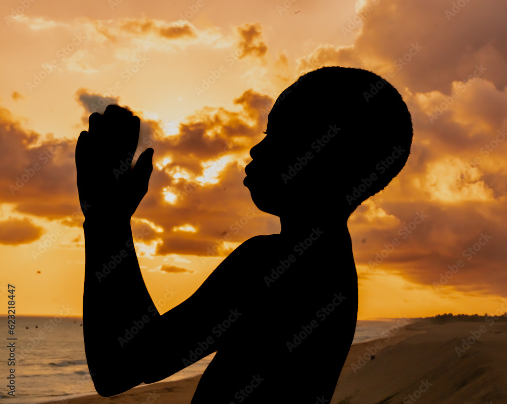 A contemplative moment frozen in time, as a young boy kneels by the ocean, his hands joined in prayer, deeply connected to the spiritual energy of the sunset.