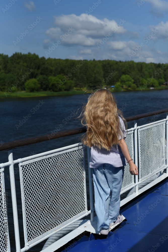 Fototapeta premium A girl with long hair stands on the deck of the ship and looks at the shore.