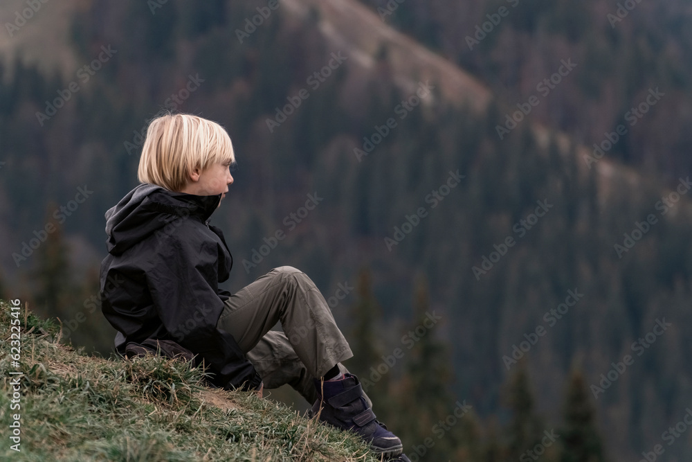Blond boy in trekking clothes on hike during break. Child is relax on the mountainside. Boy sitting on hill