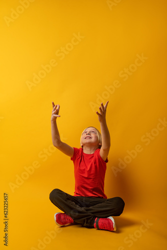 Blond schoolboy sitting cross-legged and arms up catching something on yellow background. Copy space. Vertical frame.