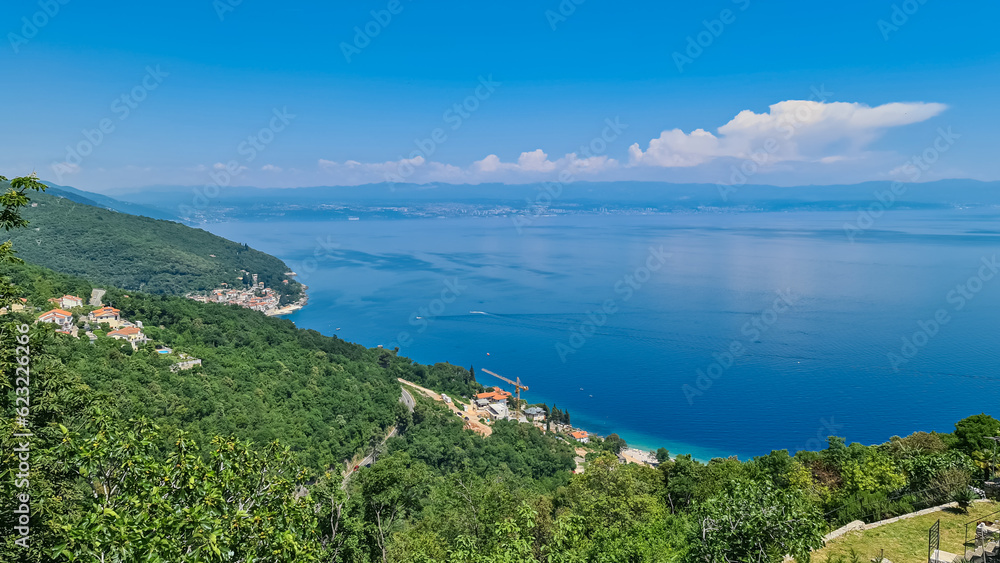 A view on the Mediterranean Sea from a viewpoint of Moscenice, Croatia. The slopes are overgrown with lush green plants. Small towns located at the sea shore. High mountains in the back. Blue sky.