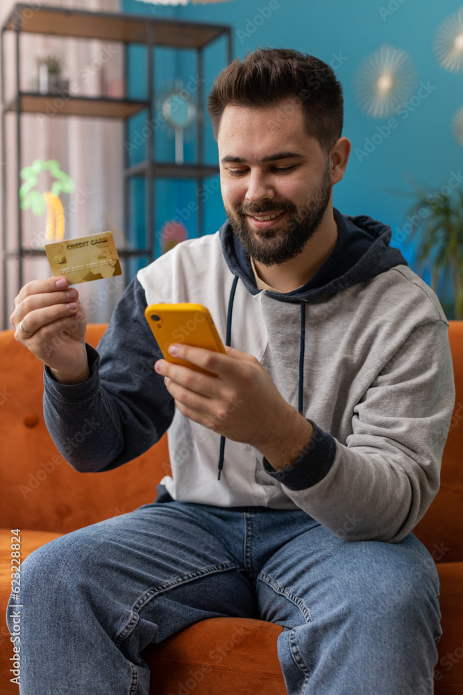 Caucasian man using credit bank card and smartphone while transferring money, purchases online shopping cashless order food delivery at home apartment indoors. Happy young guy sitting in room on couch Caucasian man using credit bank card and smartphone while transferring money, purchases online shopping cashless order food delivery at home apartment indoors. Happy young guy sitting in room on couch
