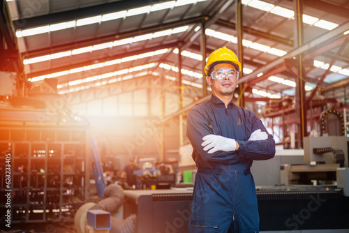 engineer man wearing bodysuit and yellow contruction helmet holding his arms with white gloves with lathe factory background with light and flare
