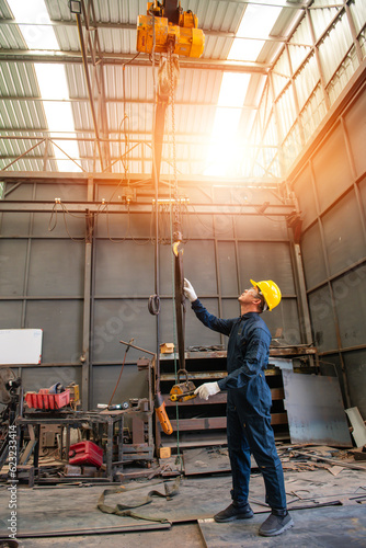 engineer man wearing bodysuit and yellow contruction helmet holding his arms with white gloves checking a hook of lifting hoist in lathe factory background with light and flare
