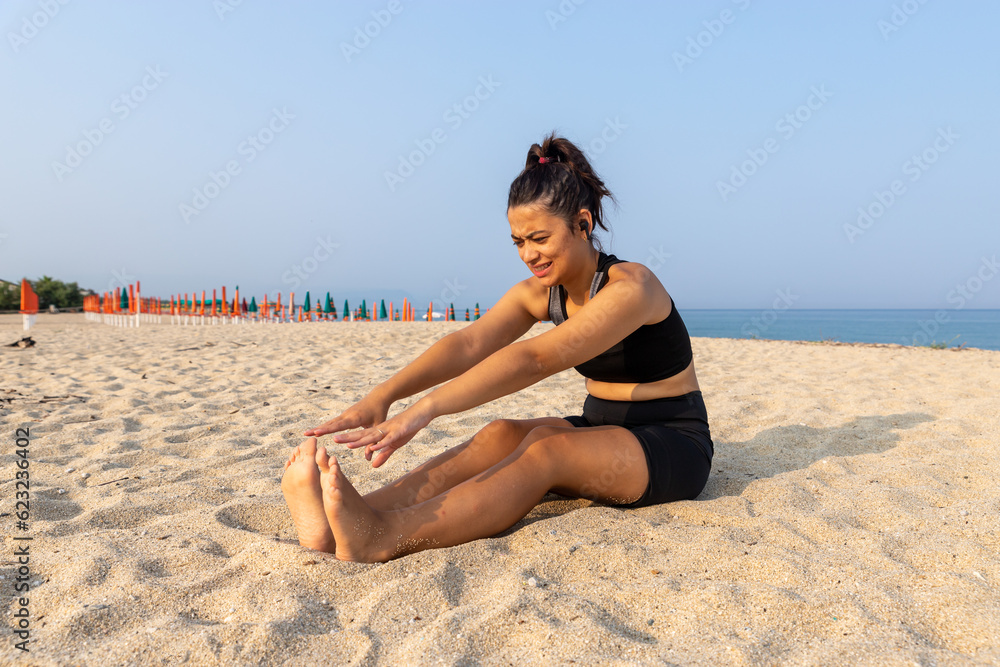 latina woman in sportswear on the beach sitting stretching with difficulty