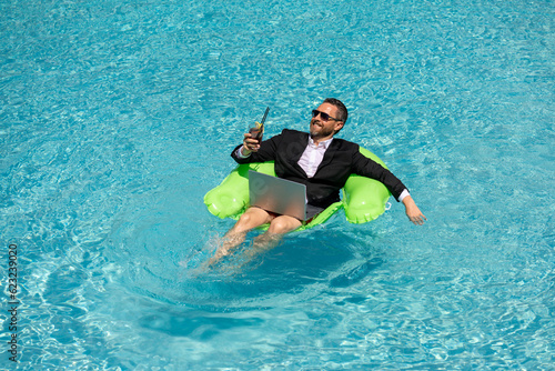 Successful businessman floating in suit in pool water. Summer vacations and travel concept. Funny business man drink summer cocktail and using laptop in swimming pool. Summer business concept. © Volodymyr
