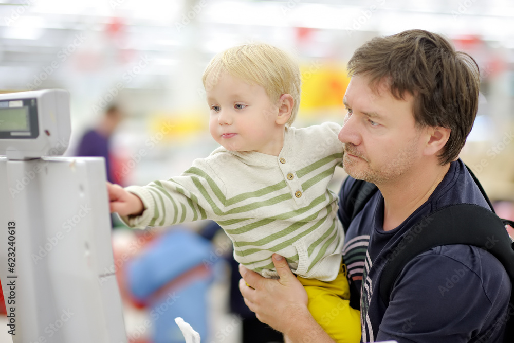 Cute toddler boy and his middle age father in a food store or a supermarket. Child helping dad to weight of groceries. Shopping with kids Cute toddler boy and his middle age father in a food store or a supermarket. Child helping dad to weight of groceries. Shopping with kids
