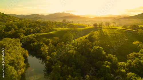 AERIAL: Morning sunbeams illuminate picturesque hilly landscape with calm river