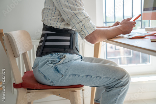 Man freelancer wearing medical orthopedic corset sitting at table working remotely from home, remote worker using lower back brace sits in front of computer, treating chronic back pain 