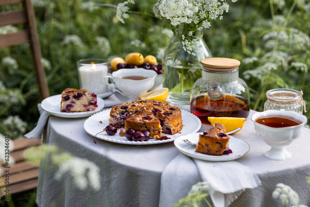 Tea time with sweet homemade cherry cake and herbal beverage in summer garden outdoors. Table covered with linen tablecloth, white porcelain cups. English style Tea time with sweet homemade cherry cake and herbal beverage in summer garden outdoors. Table covered with linen tablecloth, white porcelain cups. English style