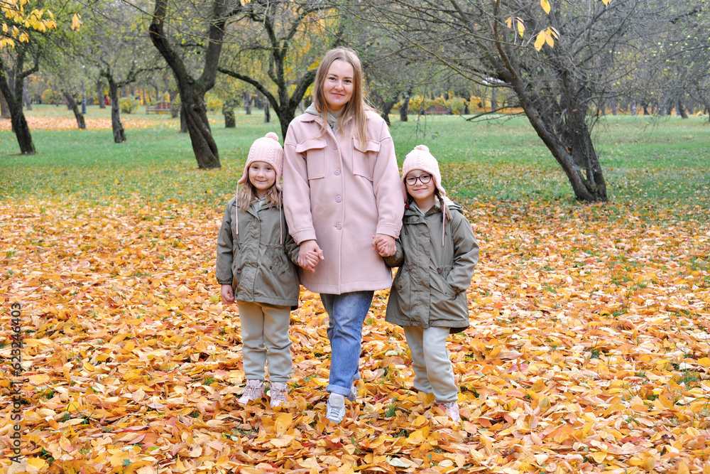 Naklejka premium Caucasian family play in autumn park, mother embrace two girls twins and smile, active weekend outdoors