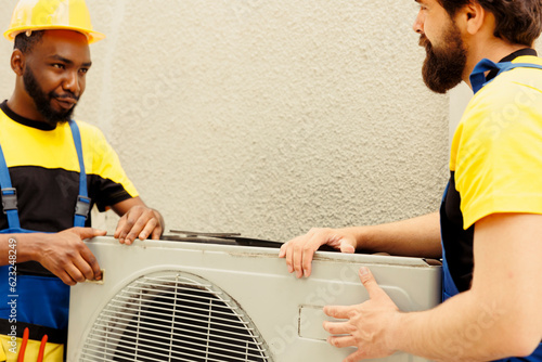 African american repairman and apprentice worker pulling apart old defective outdoor hvac system to replace it with new performant condenser after draining refrigerant and replacing ductwork photo